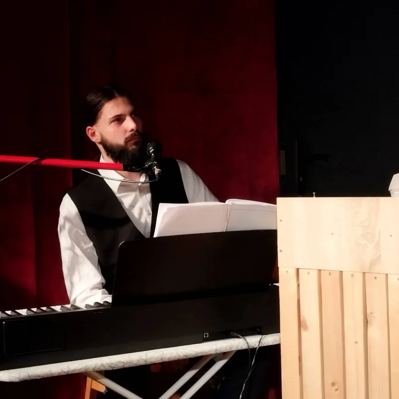 Organista w kamizelce przy klawiszach i mikrofonie, na tle czerwonej kurtyny